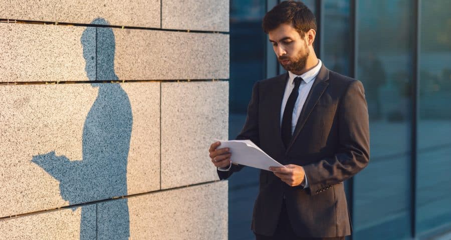 Serious young businessman in a classic suit reading advantageous business document in paper