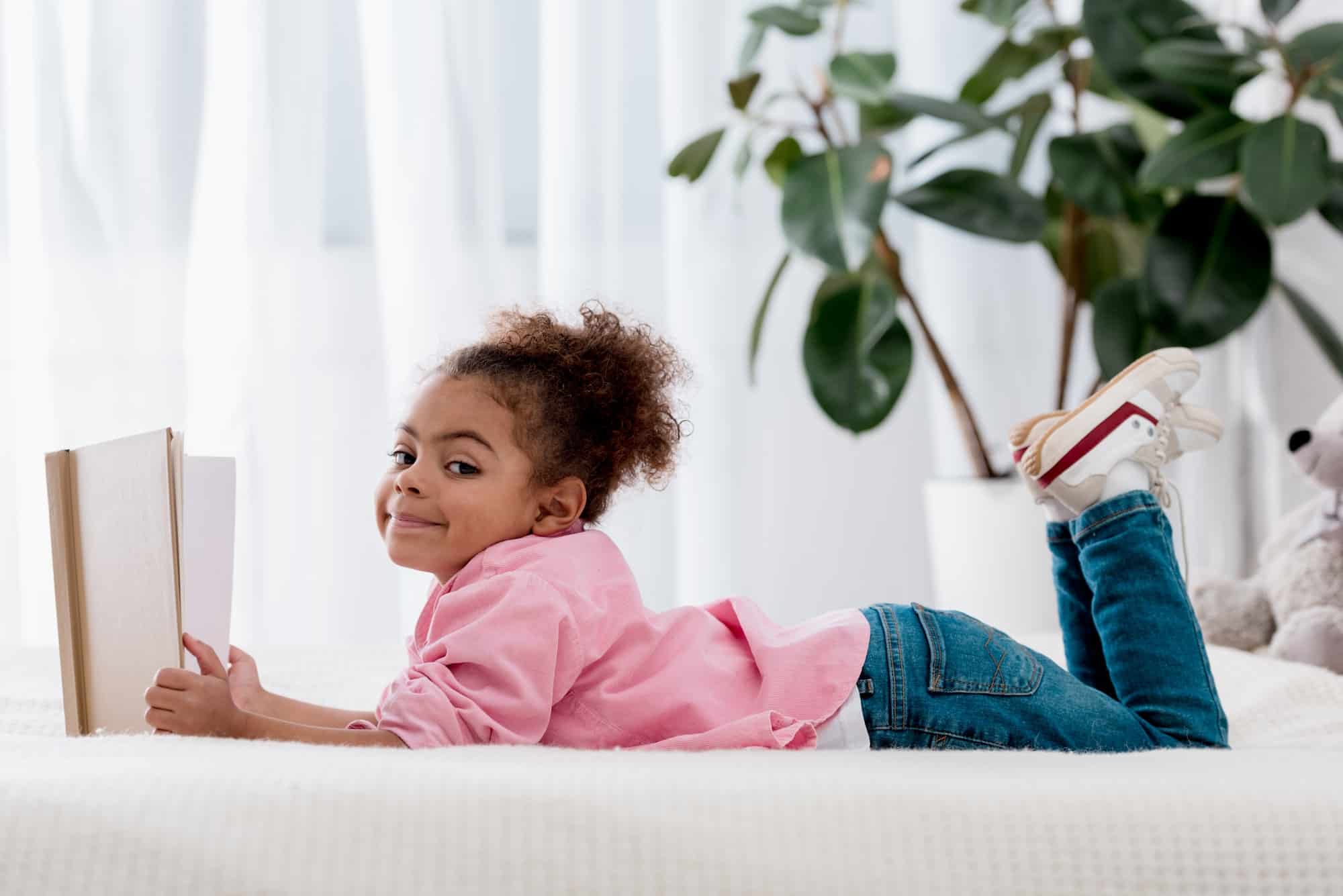 Side view of adorable African american child laying on the bed and reading the book